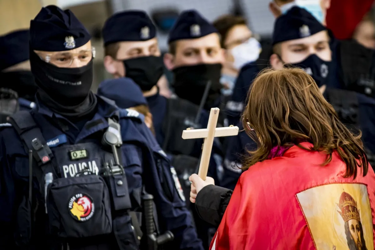 Woman in a red cape holding a wooden cross before masked police officers